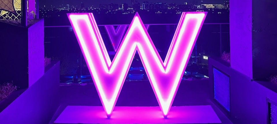 A large neon W glowing pink on a hotel rooftop at night, the city skyline behind it